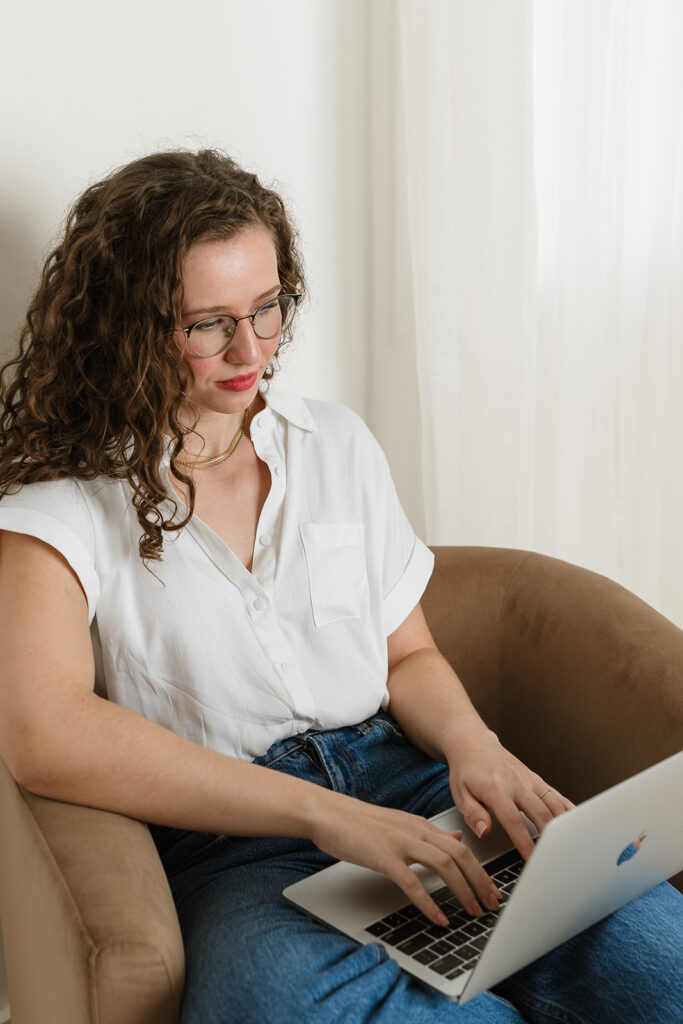 A woman with curly hair and glasses, wearing a white shirt and blue jeans, sits in a brown chair while working on a laptop.